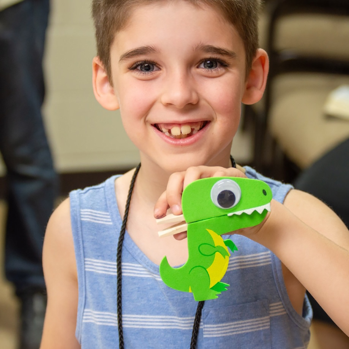 Primary boy holds up dinosaur craft from VBS 2023, Stompers & Chompers.